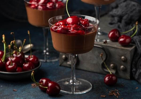 Chocolate mousse dessert topped with fresh red cherries in a coupe glass, surrounded by scattered cherries and bowls on a